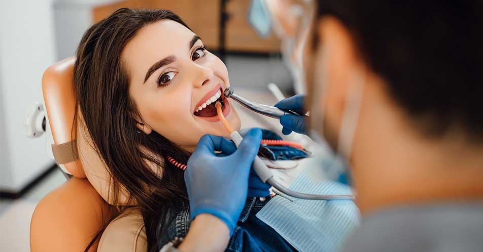 a imagem mostra uma mulher de cabelos escuros deitada em uma cadeira de consultório odontológico. Ela encara a câmera enquanto o dentista, de costas, realiza a limpeza bucal.