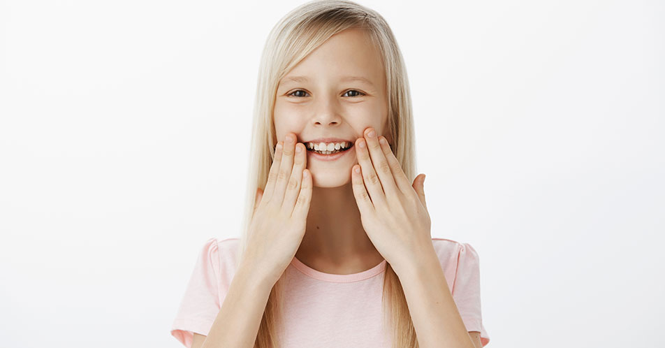 A imagem mostra uma menina, branca de cabelos loiros, longos e lisos, sorrindo. Ela veste uma camiseta rosa claro e está com as duas mãos posicionadas nas laterais do seu rosto. O fundo da imagem é branco.